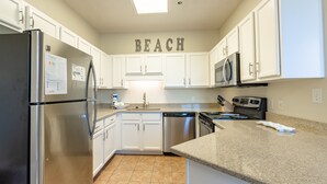 A kitchen with white cabinets, stainless steel appliances, and a 'BEACH' sign on the wall. - Phoenix East 008 by Liquid Life (Orange Beach)