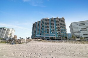 A beachfront hotel with multiple floors, a sandy beach in the foreground, and palm trees in the background.