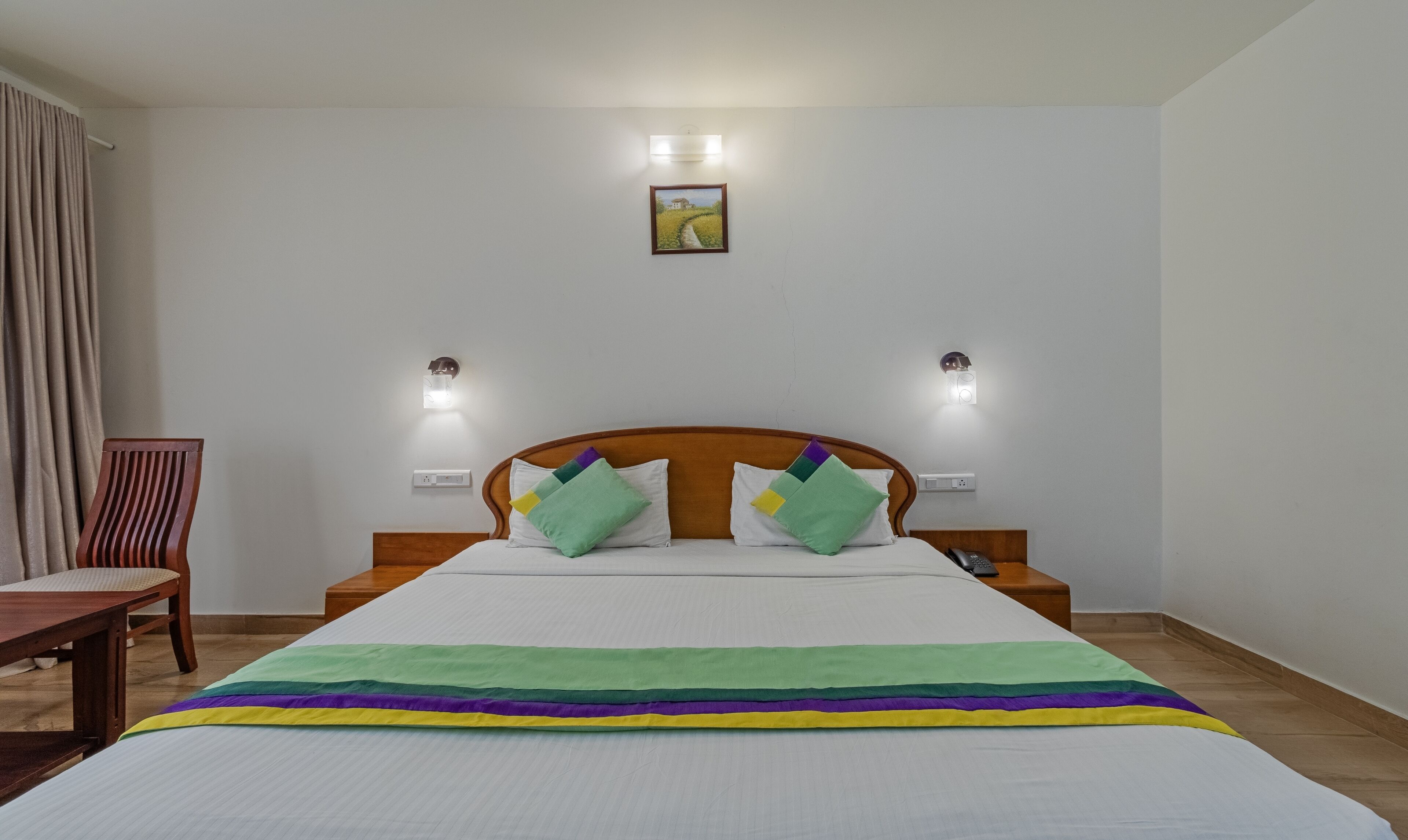 A hotel room with a wooden headboard, a bed with a rainbow-striped blanket, two green pillows, and a painting on the wall.