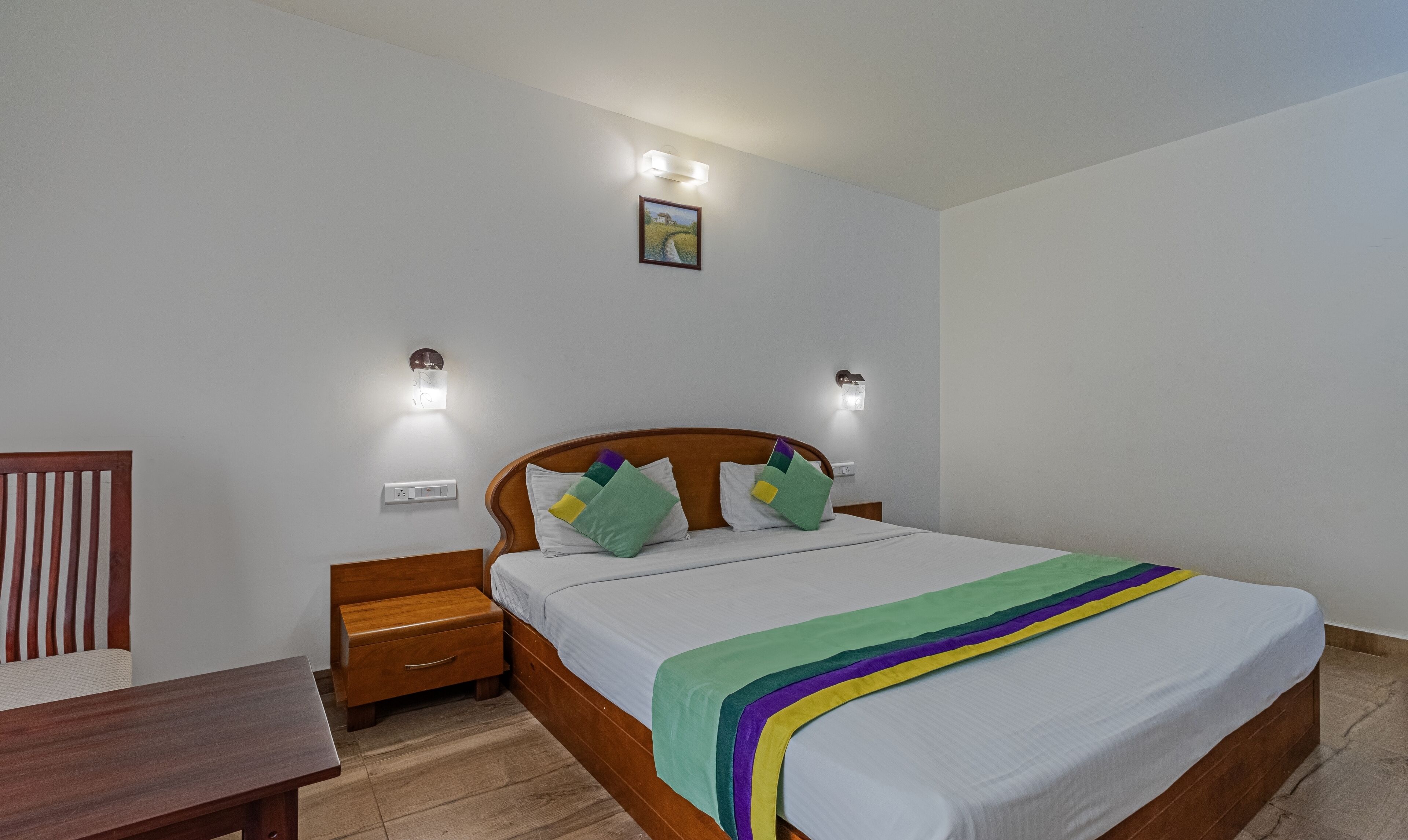 A bedroom with a wooden bed, a nightstand, a bedside table, and a wall-mounted light fixture.