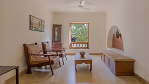 A room with a sofa, chairs, a table, a cabinet, a ceiling fan, a window with a view of greenery, and a painting on the wall. - Treebo Sadhoo Heritage (Kannur)