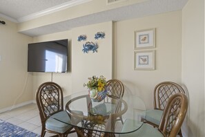 Un comedor con mesa de vidrio rodeada de sillas de mimbre, televisor de pantalla plana en la pared y decoración mural.