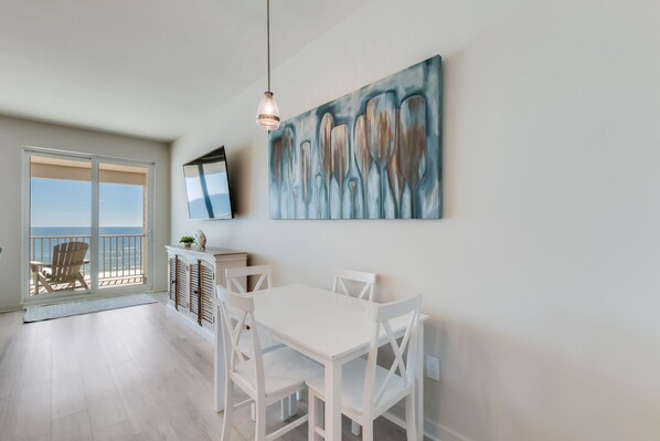 Ein Zimmer mit Esstisch, Stühlen, einem an der Wand befestigten Fernseher und Meerblick durch eine Schiebetür.