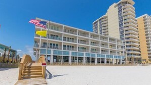 A beachfront hotel with multiple balconies, a flag, and a wooden staircase leading to the beach. - Sea Glass 406 by Liquid Life (Gulf Shores)