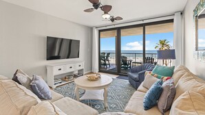 Een ruime woonkamer met een grote televisie, comfortabele zitplaatsen, een plafondventilator en een balkon met uitzicht op het strand.