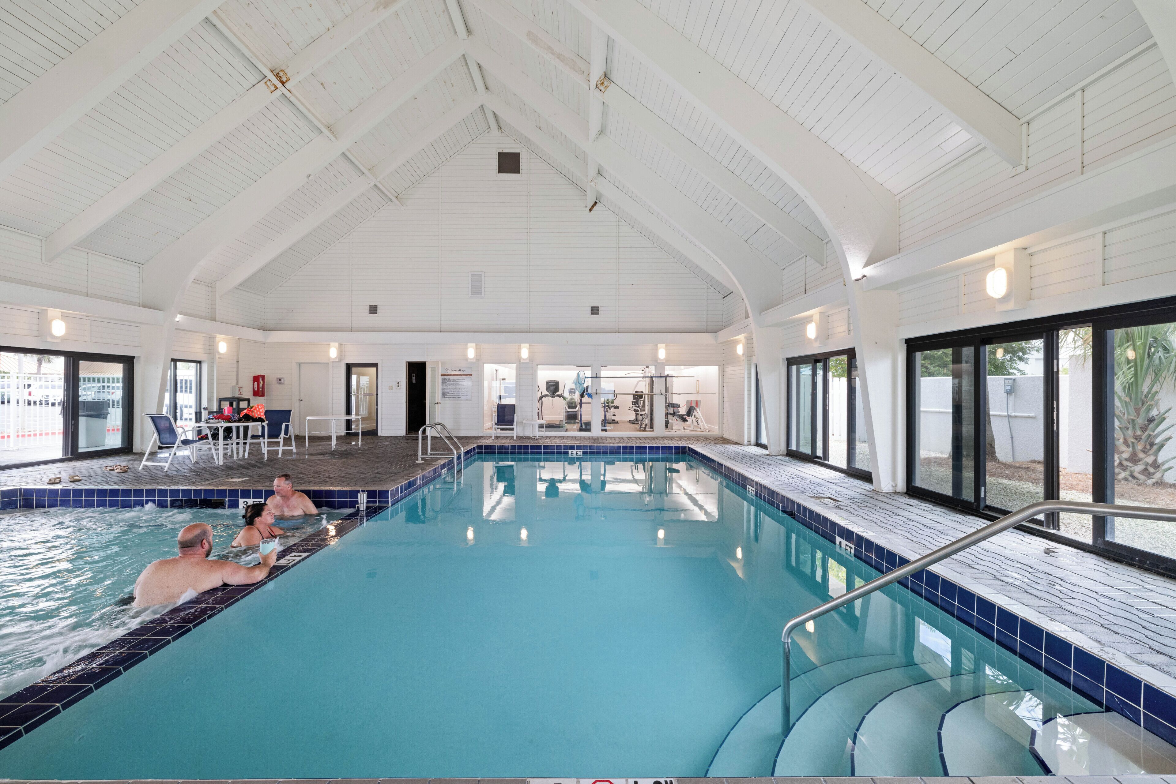 Une piscine intérieure à l’eau d’un bleu cristallin, avec un plafond blanc et une vue sur l’extérieur grâce à de grandes fenêtres.