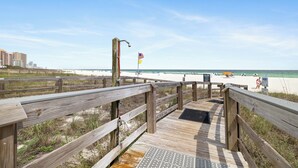 A wooden boardwalk leading to a beach with a flag, a sandy area, and buildings in the background. - Summer House 304B by Liquid Life (Orange Beach)