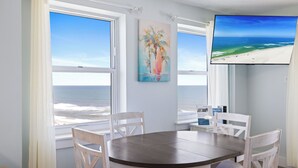 A room with a round table, white chairs, a painting, a TV, and a view of the ocean. - Tradewinds 701 by Liquid Life (Orange Beach)