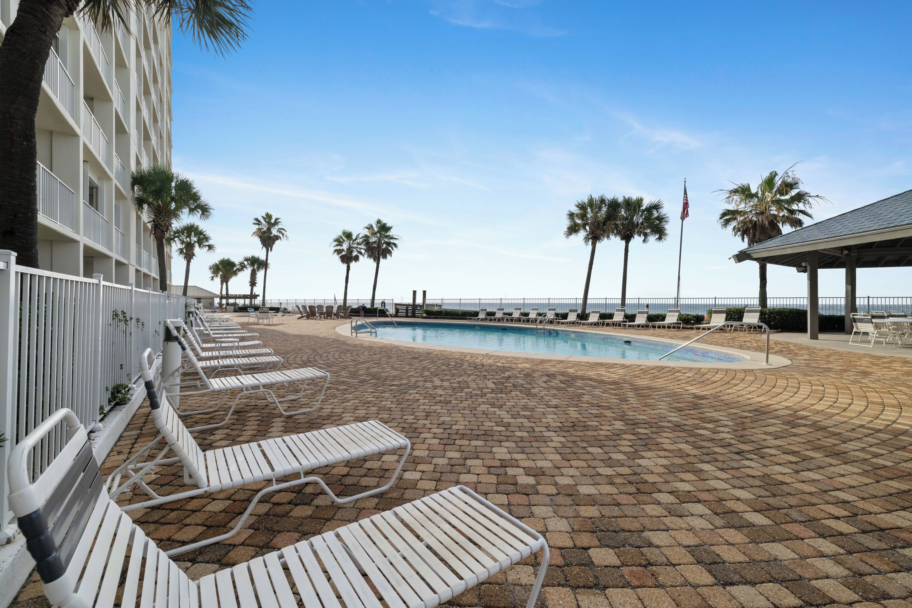 Zona de piscina con sillones, una piscina, palmeras y un edificio con balcones.