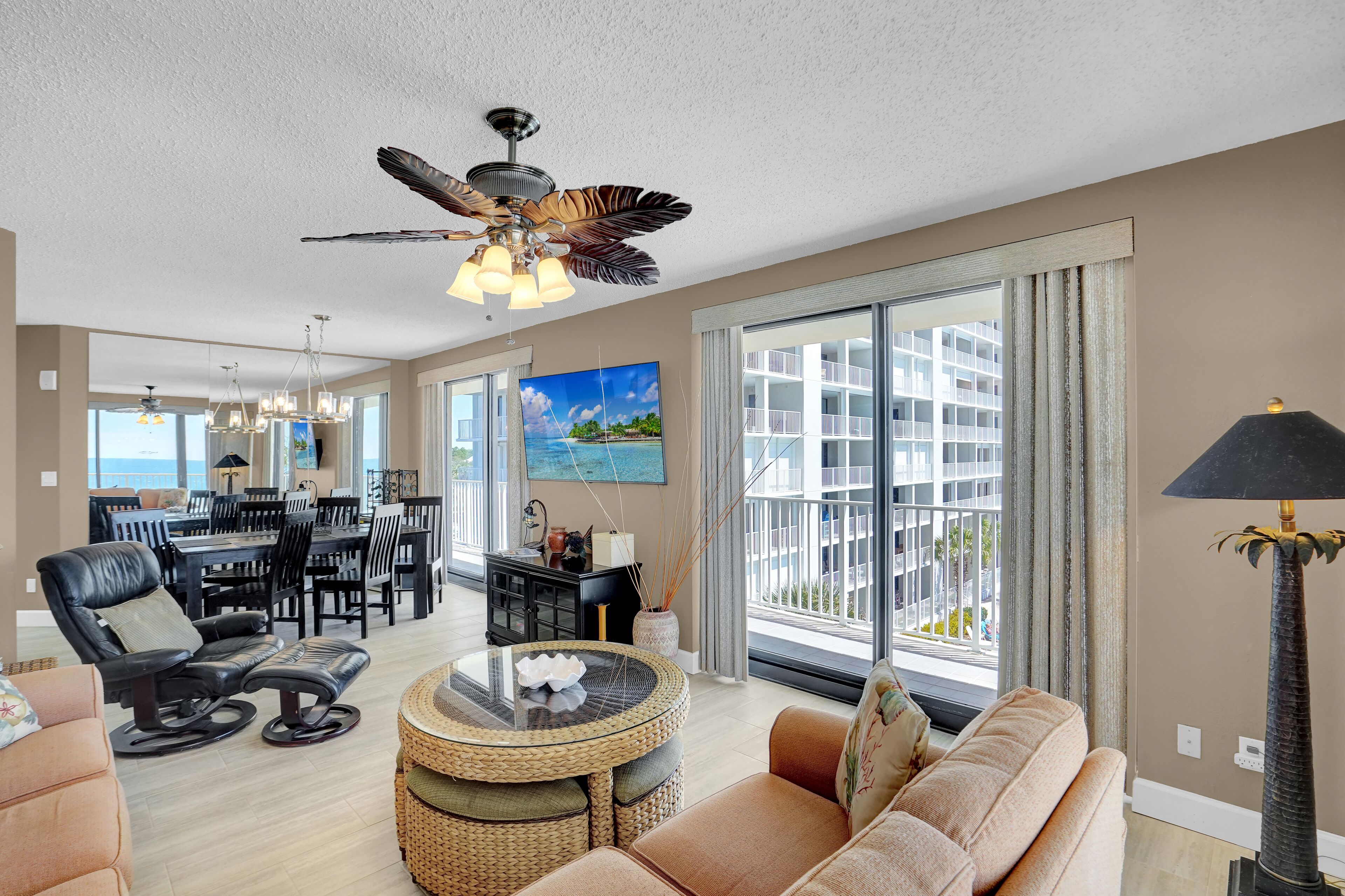 A spacious living area with a dining table, comfortable seating, a ceiling fan, and a large window offering a view of a building.
