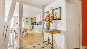 A hallway with a white door, a round gold table with a vase of flowers, and a kitchen in the background.