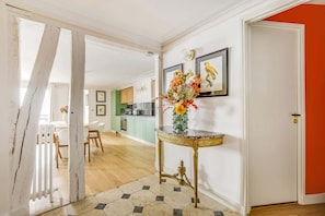Un couloir avec une porte blanche, une table ronde dorée ornée d’un vase de fleurs, et une cuisine en arrière-plan.