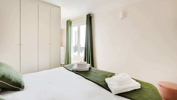A hotel room with a bed, green curtains, a view of the outdoors, and a white wardrobe.