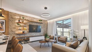 A modern living room with a large flat-screen TV, comfortable seating, a dining area, and a view through large windows. - Family 4BR Home w/ Pool at Storey Lake (Kissimmee)