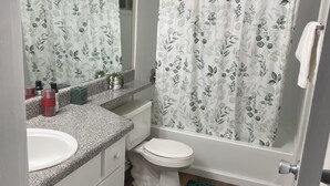 Ein Badezimmer mit weißem Waschbecken, Toilette und einer Duschvorhang mit floralem Muster. Auf dem Boden liegen grüne Teppiche und über dem Waschbecken hängt ein Spiegel.