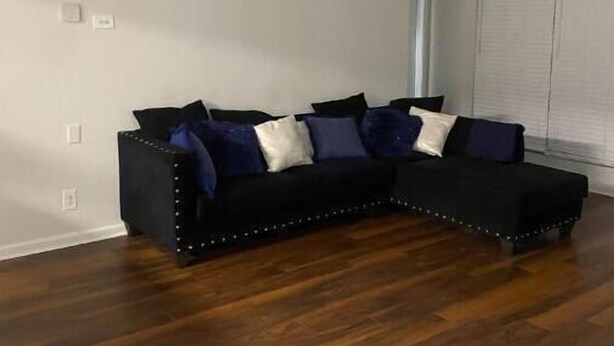 Ein Eckwohnzimmer mit einem dunkelgrauen Ecksofa, weißen und dunklen Kissen sowie Holzdielen.