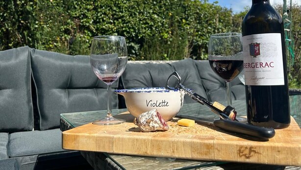 A bottle of Bergerac wine, two glasses, a bowl, and a cheese on a wooden table outdoors with a hedge and sky in the background. - Cosy rural 5 bedroom home with games room, pet friendly, garden, bbq! (Ger)