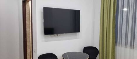 A small room with a flat-screen TV mounted on the wall, two black chairs, a small round table, and a green curtain.