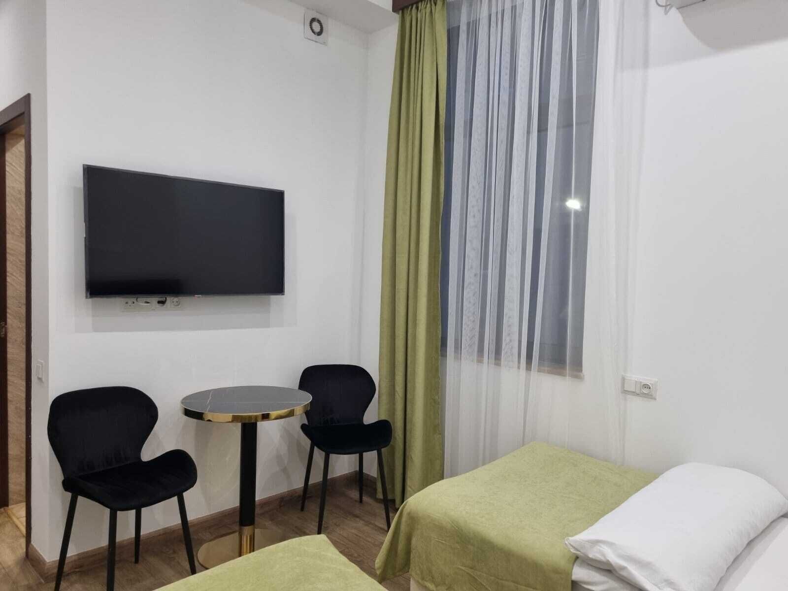 A hotel room with a TV mounted on the wall, two black chairs around a small round table, and two beds with green and white bedding.