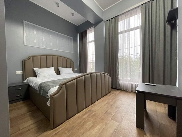 A bedroom with a bed, a bedside table, a desk, and large windows with curtains.