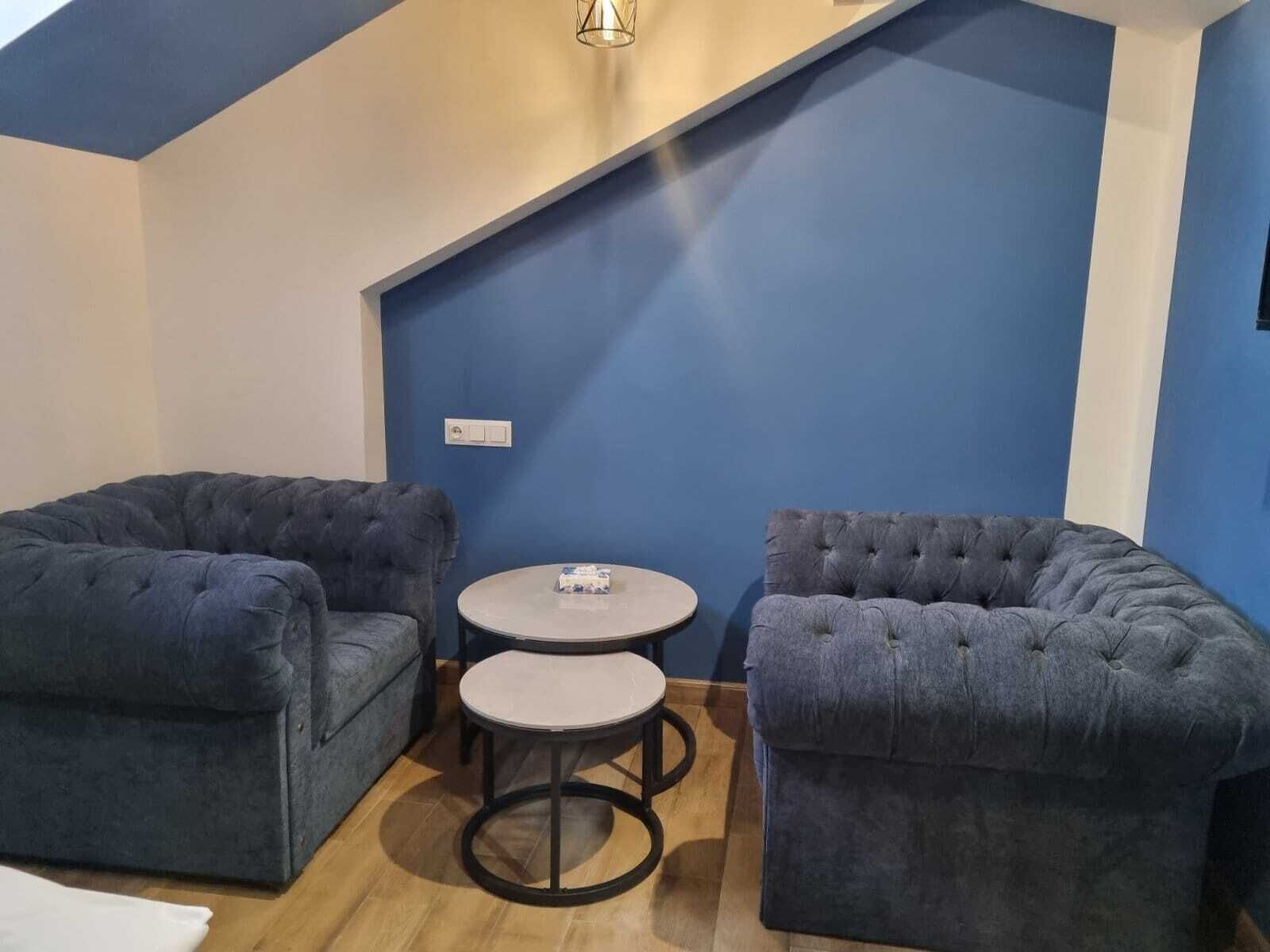 Een kamer met twee ruime fauteuils, een kleine ronde tafel, een lamp aan het plafond en een blauwe accentmuur.