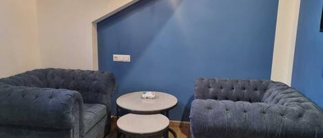 Een kamer met twee ruime fauteuils, een kleine ronde tafel, een lamp aan het plafond en een blauwe accentmuur.