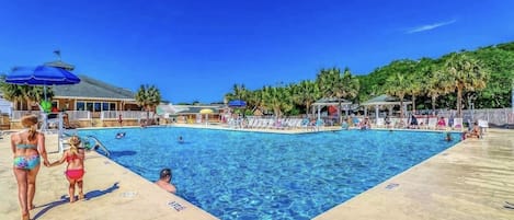 A swimming pool with lounge chairs and umbrellas, surrounded by palm trees and a building with a sloped roof.