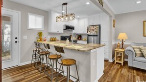 A modern kitchen with white cabinets, a central island with bar stools, a hanging light fixture, and a small side table with a lamp. - N39 Ocean Lakes 2 Bd 2 bath golf cart included! (Myrtle Beach)