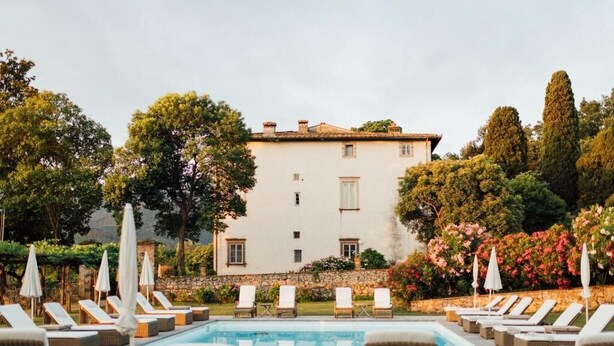 A poolside area with lounge chairs and umbrellas, a white building with a tiled roof, and lush greenery in the background. - Buonvisi B&B (Lucca)