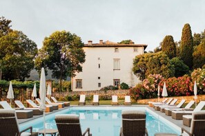 A poolside area with lounge chairs and umbrellas, a white building with a tiled roof, and lush greenery in the background. - Buonvisi B&B (Lucca)