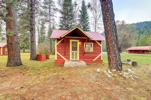Una pequeña cabaña roja con una puerta amarilla, rodeada de árboles y un espacio cubierto de césped.