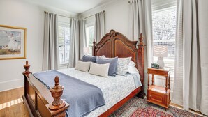 A bedroom with a wooden bed, a nightstand with a lamp, and a view of the city through the window. - Historic Charnwood Guest House- 1 Mile to Downtown (Tyler)