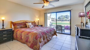 A bedroom with a bed, a ceiling fan, a nightstand, a dresser, and a view of the outdoors. - Palm Desert Condo w/ Patio & Golf Course Views! (Palm Desert)