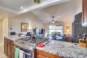 A kitchen with a large island, stove, and granite countertop. There is a dining area with a table and chairs, and a view of the outdoors through a window. - Palm Desert Condo w/ Patio & Golf Course Views! (Palm Desert)