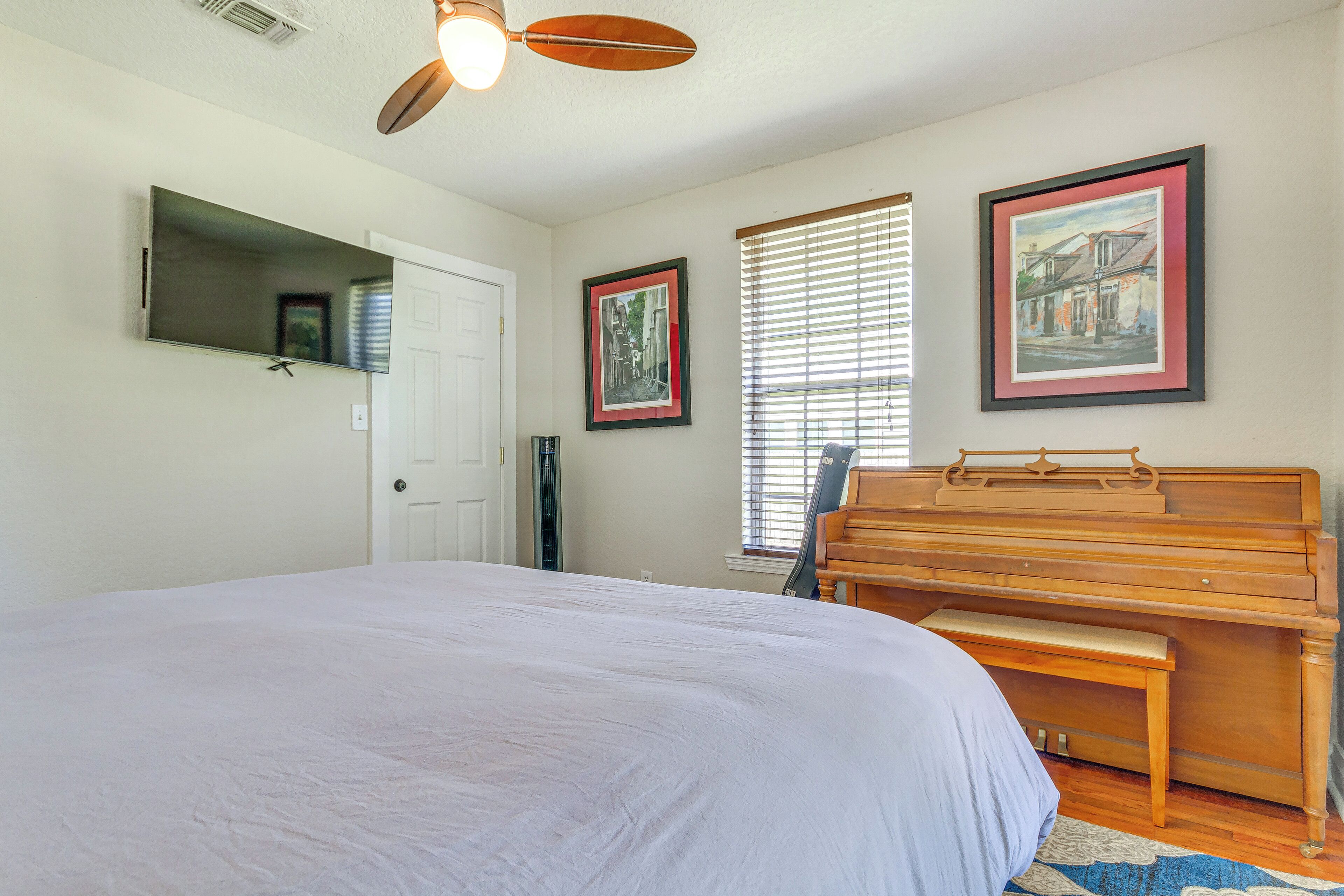 A bedroom with a large bed, a wooden piano, a ceiling fan, and two framed pictures on the wall.