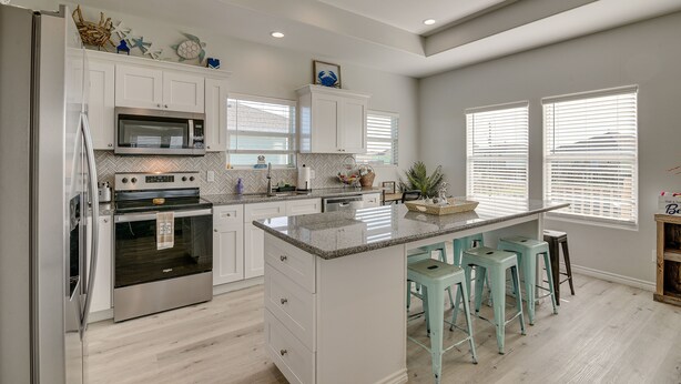 A modern kitchen with white cabinets, a stainless steel refrigerator, a built-in oven, and a granite countertop. There are three bar stools and a window with blinds. - Crystal Beach Home w/ Covered Patio: Pets Welcome! (Port Bolivar)