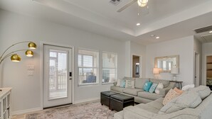 A living room with a gray sectional sofa, a coffee table, and a patterned rug. There are two windows with blinds, a door, and a ceiling fan. - Crystal Beach Home w/ Covered Patio: Pets Welcome! (Port Bolivar)