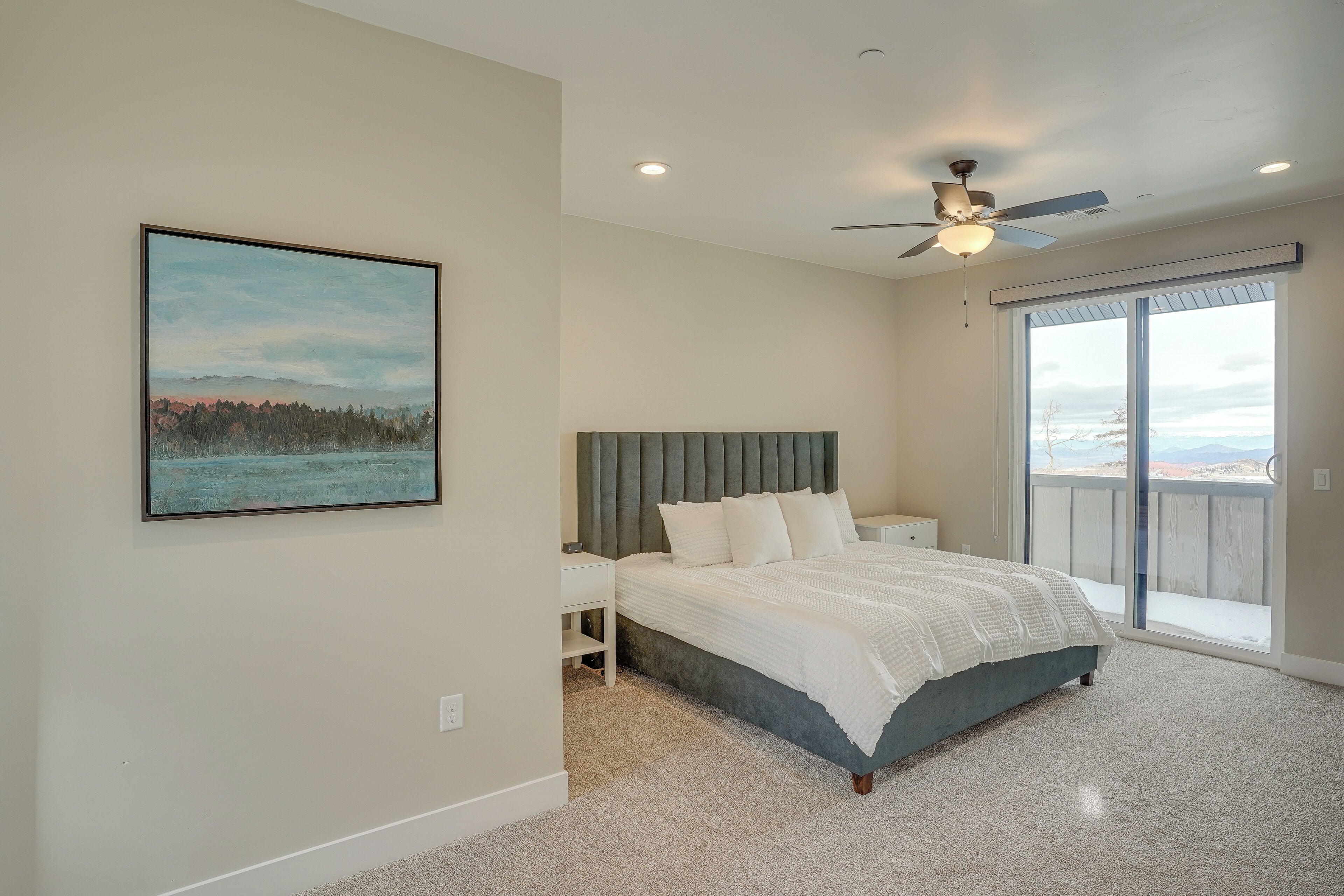 A bedroom with a bed, a ceiling fan, a large window, and a painting on the wall.