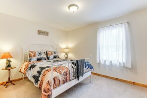 A bedroom with a white metal bed frame, a patterned bedspread, a bedside lamp, a small table with a lamp, a window with sheer curtains, and a 'Together' sign on the wall.