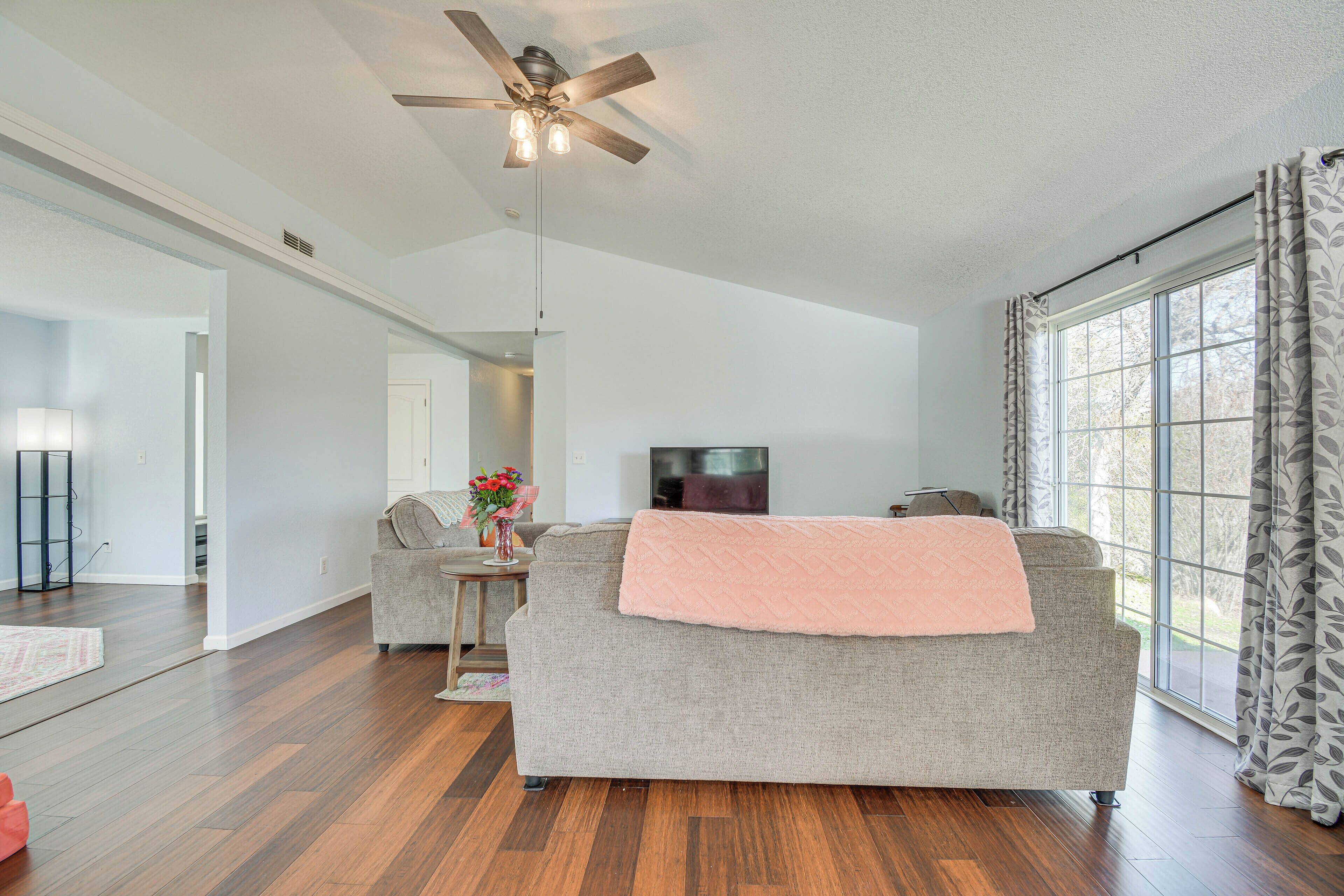 A spacious living room with hardwood flooring, a grey sofa, a ceiling fan, and a large window with curtains.
