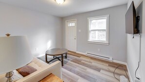 A room with a sofa, a round table, a lamp, a TV mounted on the wall, and a window with blinds.