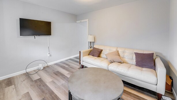 A living room with a flat-screen TV mounted on the wall, a beige leather sofa with cushions, a round ottoman, and a lamp on a side table.