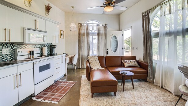 A modern kitchen with white cabinets, a microwave, and a stove. There is a brown leather sofa and a small table in the living area. A ceiling fan is visible, and the room has large windows with curtains. - Cozy Alpine Cottage Near Casinos & Trails! (Alpine)