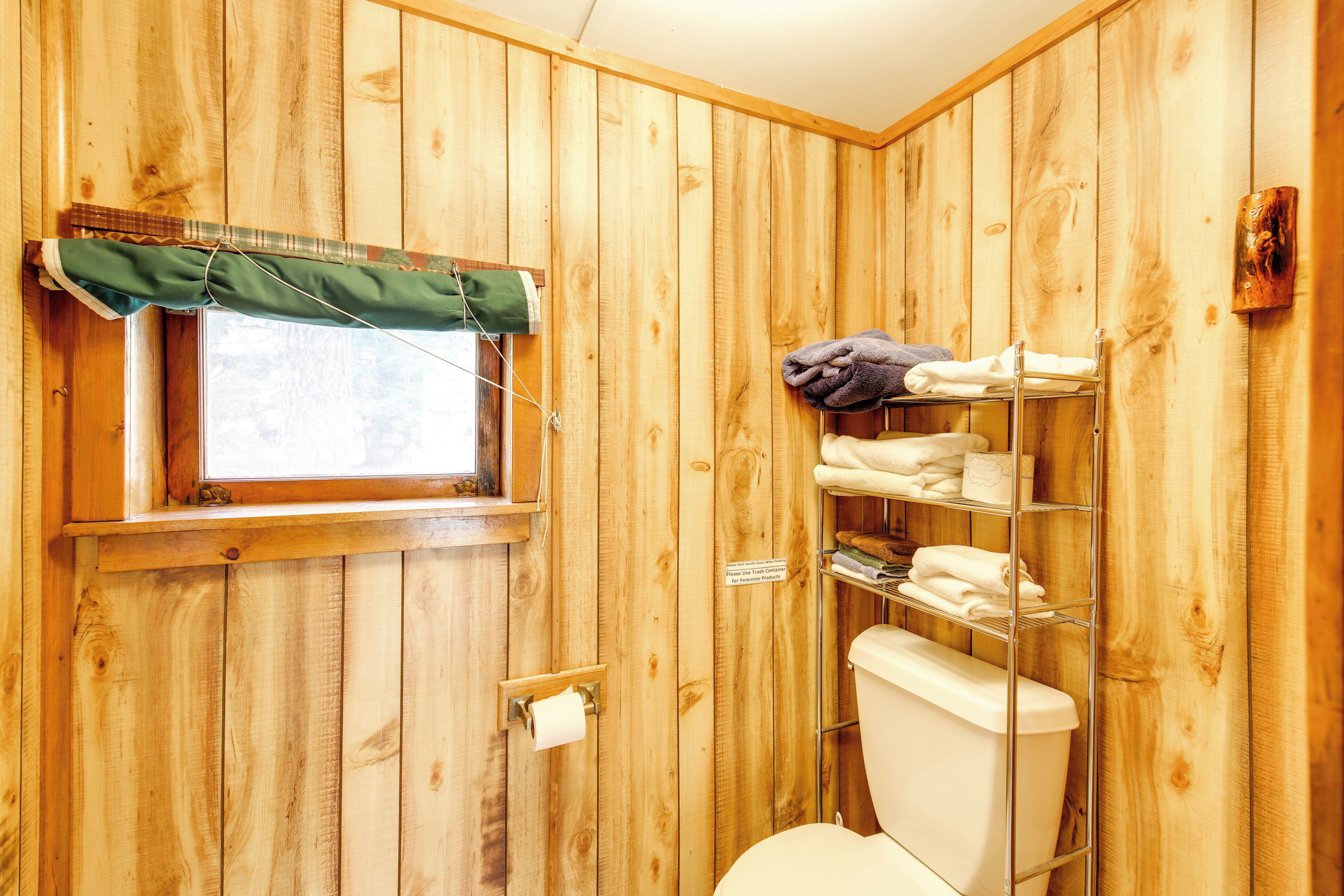 Une salle de bain aux murs lambrissés, équipée de toilettes, d’une étagère avec des serviettes et d’une fenêtre ornée d’un rideau vert.