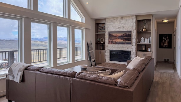 A spacious living room with a large brown leather sofa, a stone fireplace, and multiple windows offering a view of the landscape. - Great for Groups! Luxury Mtn-view Granby Home (Granby)