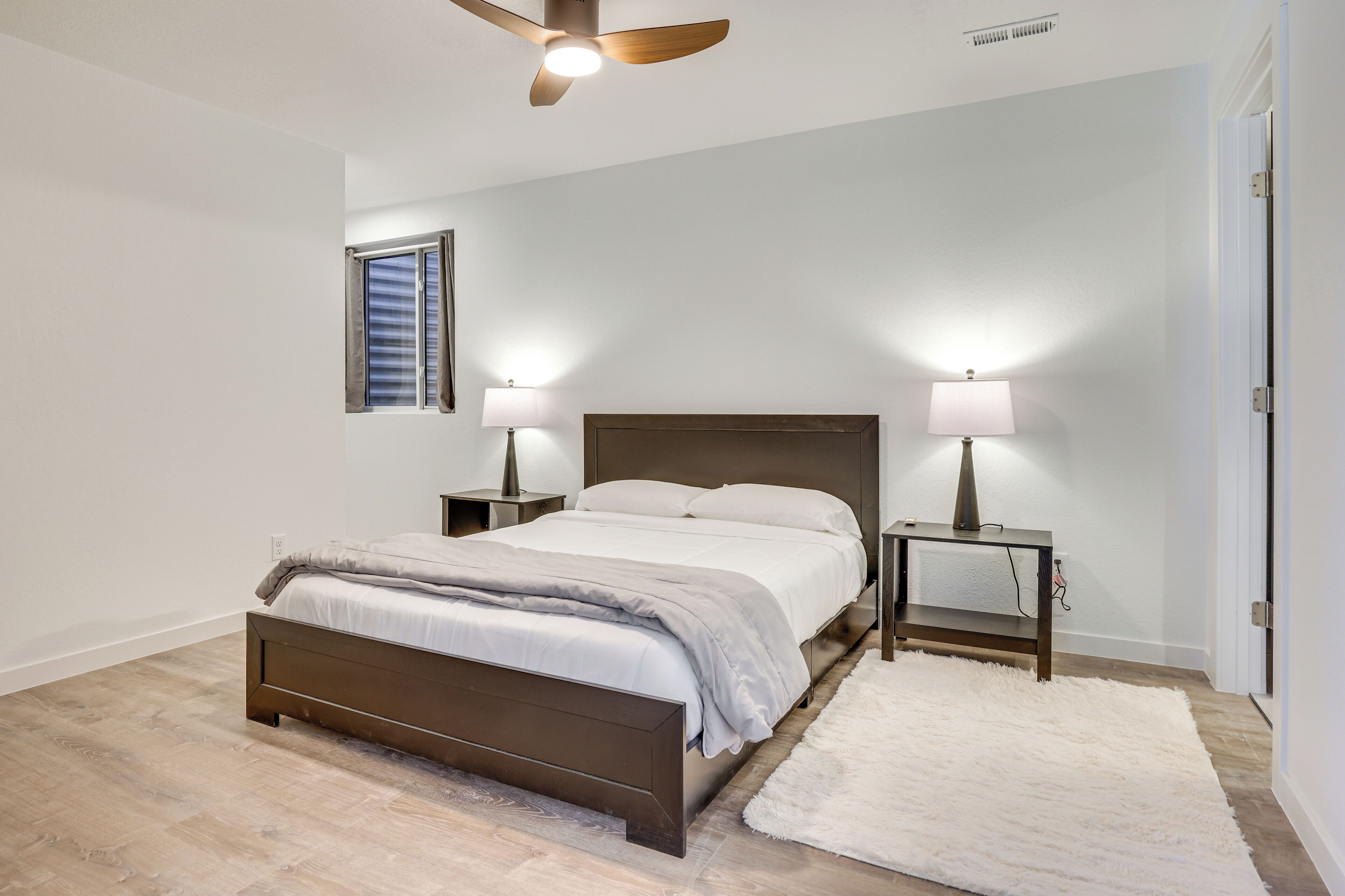 Un dormitorio con cama, dos mesitas de noche con lámparas, un ventilador de techo, una ventana con persianas y una alfombra blanca.