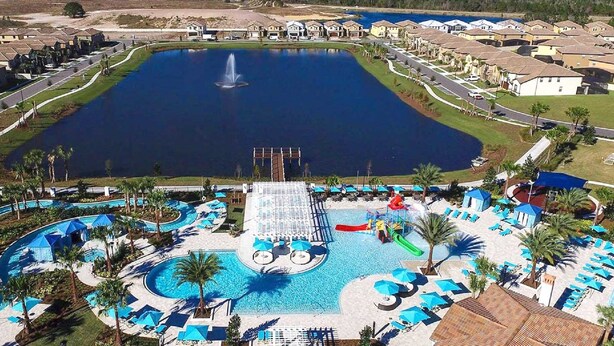 Una gran piscina con una fuente, rodeada por una zona de resort con varios edificios y una cancha de tenis.
