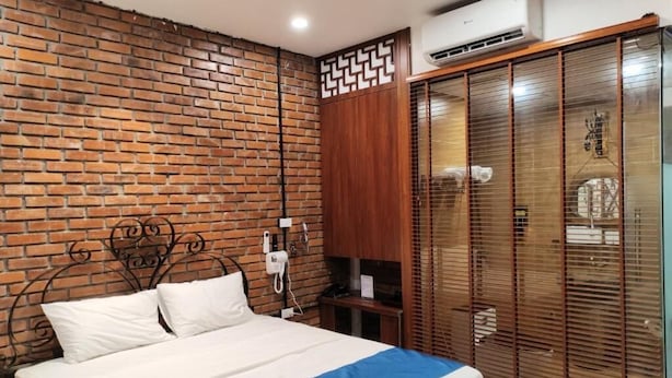 A bedroom with a brick wall, a bed with a blue and white bedspread, a wooden wardrobe, and a window with blinds. - Moc An by Bay Hotel (Hanoi)