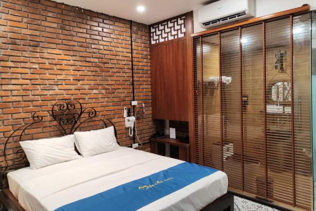 A bedroom with a brick wall, a bed with a blue and white bedspread, a wooden wardrobe, and a window with blinds.