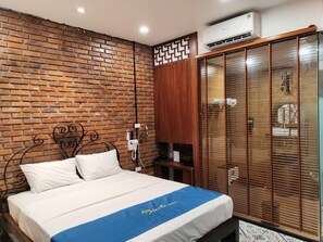 A bedroom with a brick wall, a bed with a blue and white bedspread, a wooden wardrobe, and a window with blinds. - Moc An by Bay Hotel (Hanoi)